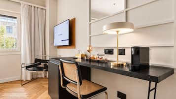 Una habitación de hotel moderna con piso de madera oscura, un escritorio negro, una silla tejida, una lámpara de pie y un televisor de pantalla plana montado en la pared.