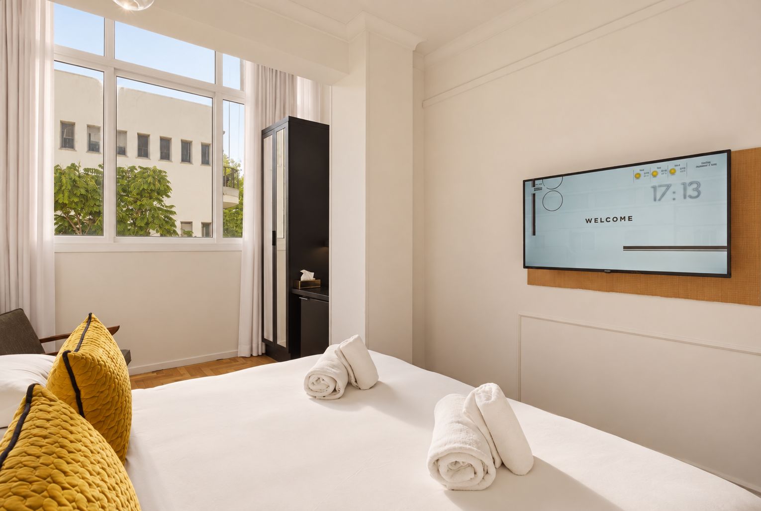 Habitación de hotel con una cama, un televisor mostrando la hora y un mensaje de bienvenida, una ventana con cortinas y vistas a edificios.