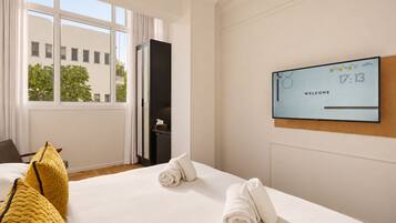 Habitación de hotel con una cama, un televisor mostrando la hora y un mensaje de bienvenida, una ventana con cortinas y vistas a edificios.