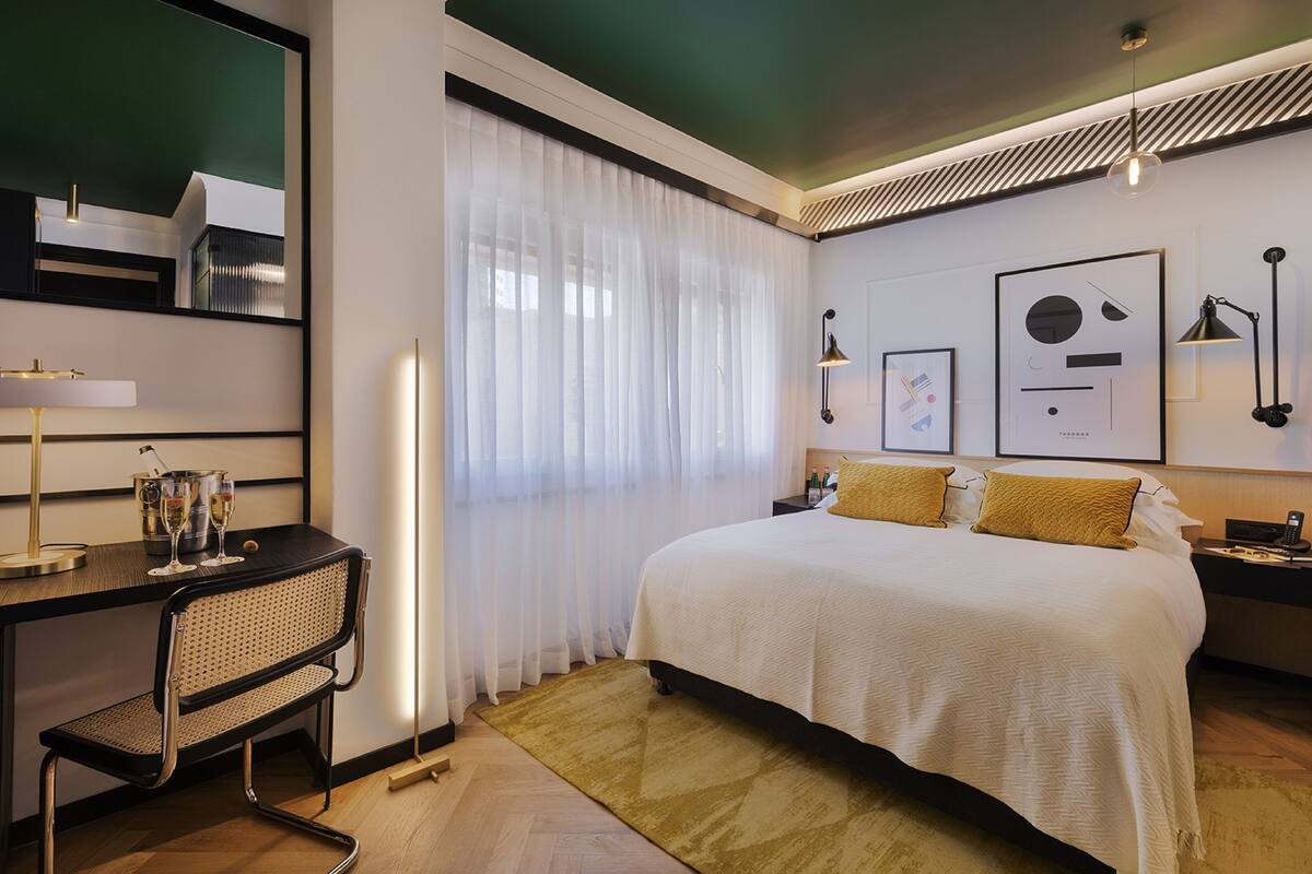 A modern hotel room with a large bed, a desk with a chair, a mirror, and artwork on the walls.