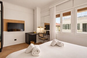 Habitación de hotel con cama, escritorio, silla, televisor y ventana con vista a edificios.