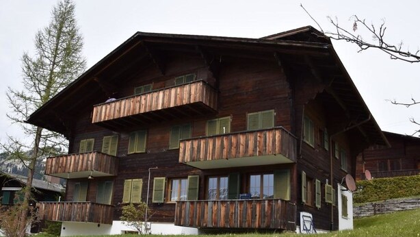Eine mehrstöckige Holzhütte mit Balkonen, eingebettet in eine grüne Wiese mit Bäumen im Hintergrund.