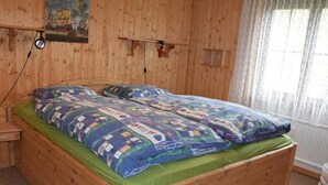 Ein hölzernes Bett mit grüner Matratze und gemusterten Betttextilien, ein Nachttisch aus Holz, eine wandmontierte Lampe und ein Fenster mit durchsichtigen Vorhängen.