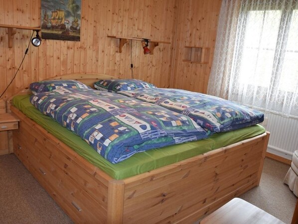A wooden bed with a green mattress and patterned bedding, a wooden nightstand, a wall-mounted lamp, and a window with sheer curtains.