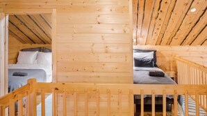 Interior de una cabaña de madera con una cama en el altillo y una cama baja, vigas de madera en el techo y una baranda de madera en primer plano.
