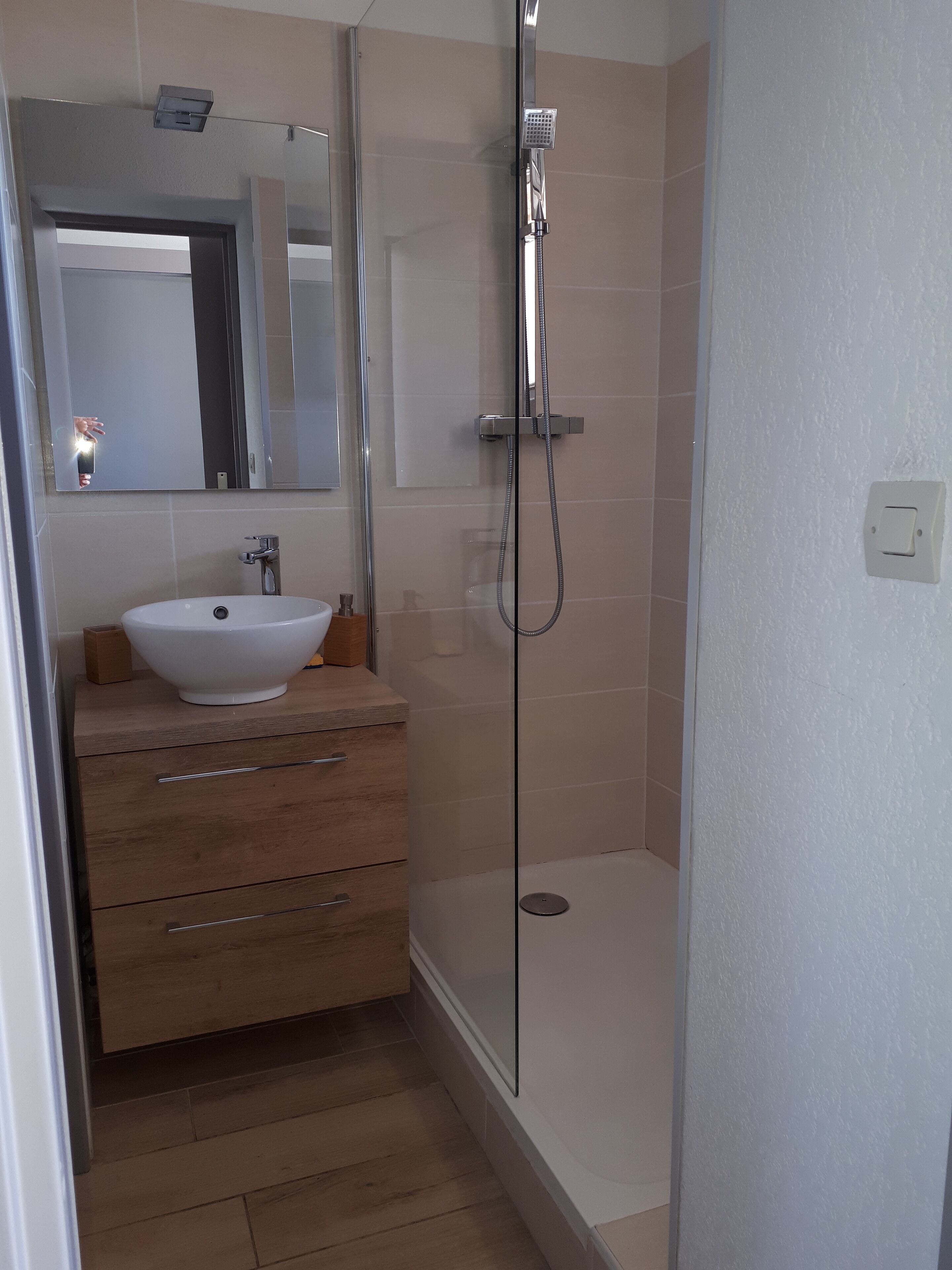 Een badkamer met een witte wastafel, houten opbergmeubel en een douche met glazen wanden.