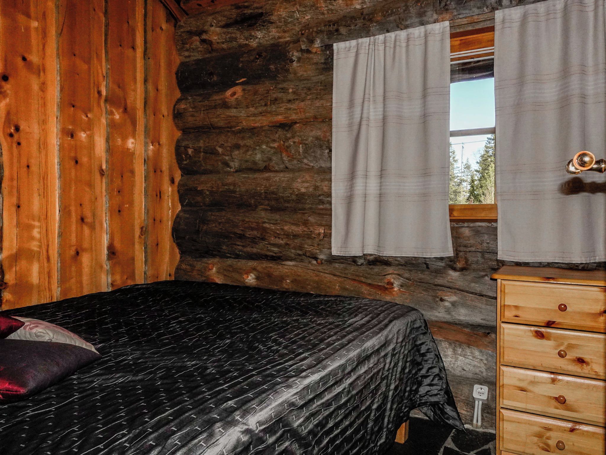 Una habitación tipo cabaña de troncos con una cama, un aparador de madera y una ventana con cortinas.