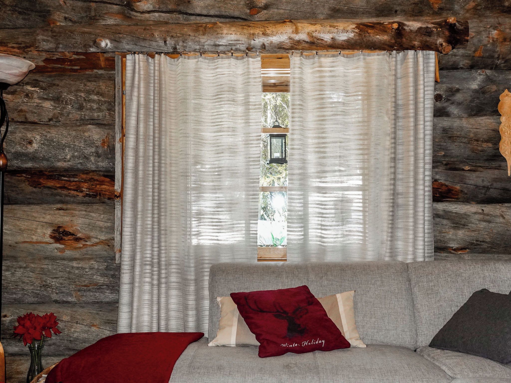Interior de una cabaña de troncos con una cama, una ventana con cortinas y un techo rústico de madera.