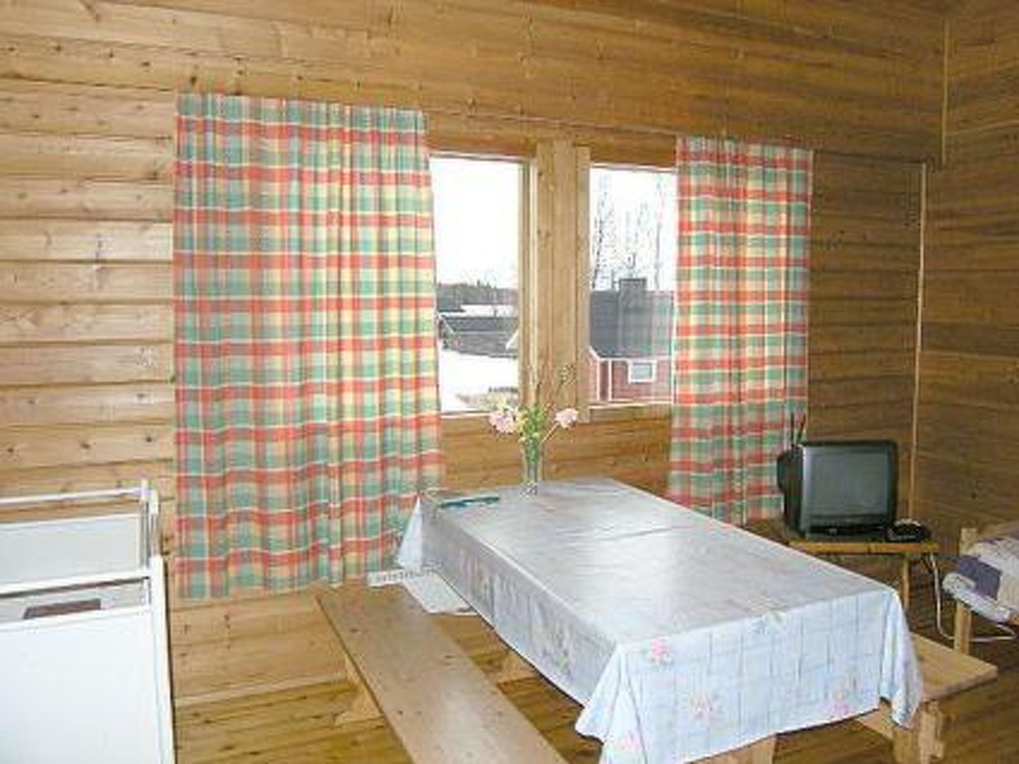 Eine Holzhütte mit einem Tisch, der mit einer Tischdecke bedeckt ist, einem Fernseher und karierten Vorhängen.