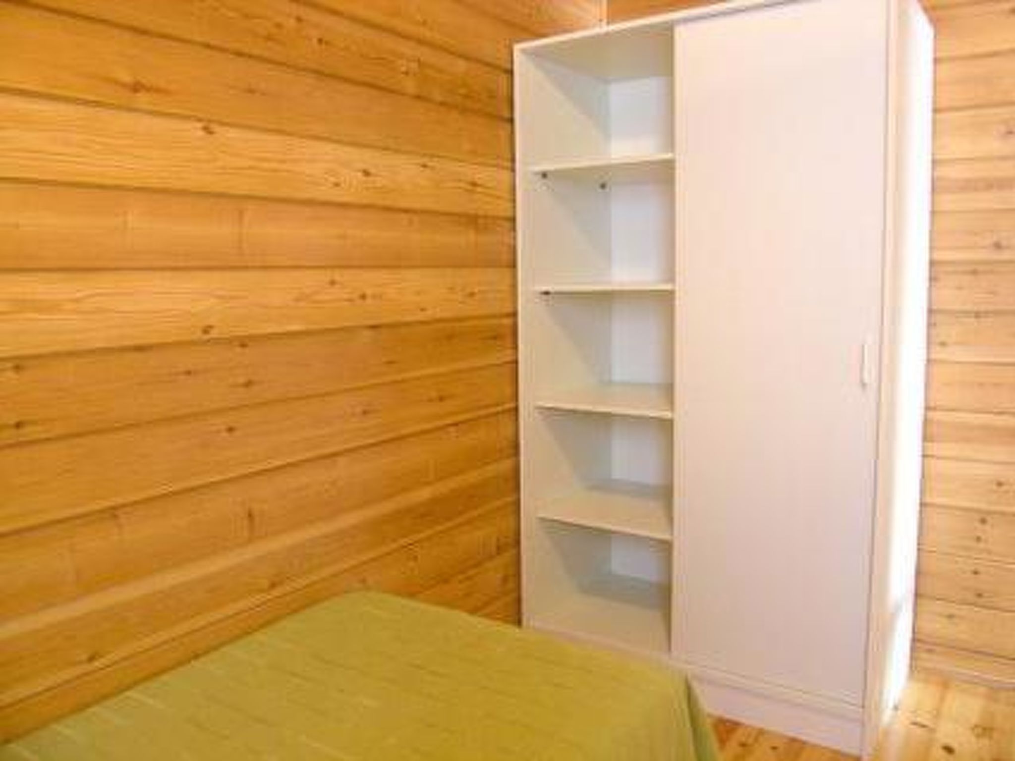 A room with wooden walls, a white wardrobe with multiple shelves, and a green bedspread.