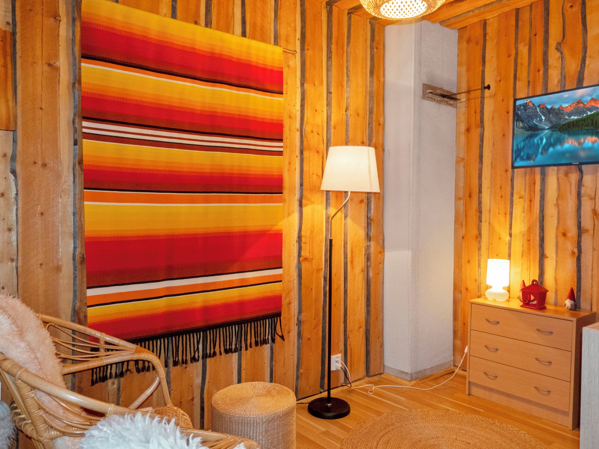 Une pièce avec une grande couverture à rayures, un lampadaire, un fauteuil en osier, un petit pouf et une commode sur laquelle est posée une télévision au mur.
