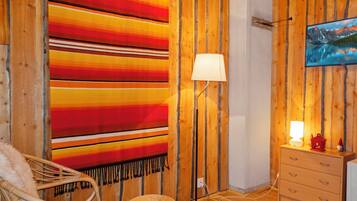A room with a large striped blanket, a floor lamp, a wicker chair, a small ottoman, and a dresser with a television on the wall.