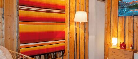 A room with a large striped blanket, a floor lamp, a wicker chair, a small ottoman, and a dresser with a television on the wall.