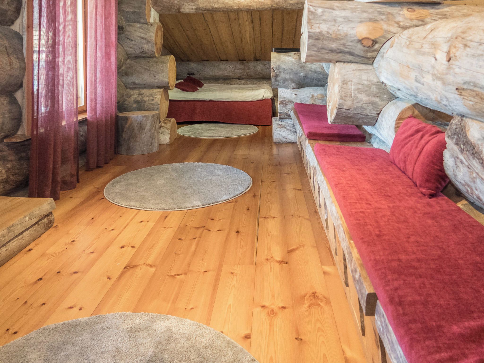Interior de una cabaña de troncos con piso de madera, un banco rojo y una cama pequeña con una colcha roja.