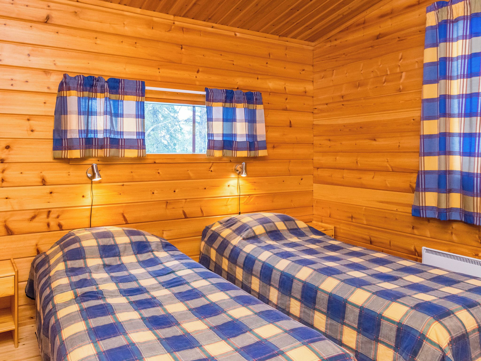 Una cabaña con dos camas, paredes de madera, una ventana con cortinas y una pequeña mesa.