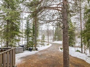 Lumipeitteinen metsäpolku johtaa puiseen mökkiin, jonka lähellä on metallinen lumilapio.