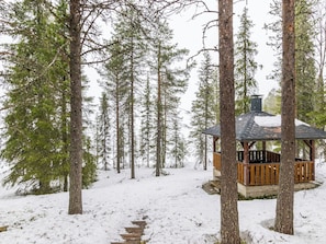Lumisessa metsässä sijaitseva puinen mökki, jonka ympärillä kohoaa korkeita, ikivihreitä puita.