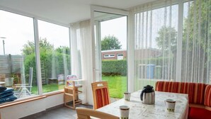 Ein sonnendurchfluteter Essbereich mit Tisch für zwei Personen, einem roten Sofa und Blick auf einen Garten durch große Fenster.