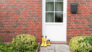 Ein rotes Backhaus mit weißer Tür, Hausnummer 23, und einem kleinen Garten mit gelben Stiefeln und grünen Büschen.