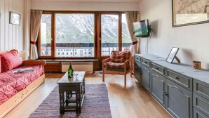 A cozy living room with a sofa, a coffee table, and a chair. There is a TV mounted on the wall and a window with a view of mountains. - Le Bec Rouge (Le Lac) by Interhome (Tignes)