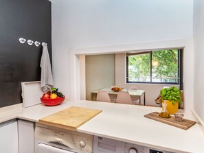 A compact kitchen with a wooden countertop, a red bowl of fruit, a cutting board, a sink, and a window with a view of greenery.