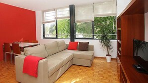 Un salon moderne avec un canapé d’angle beige, une table à manger avec des chaises, un téléviseur et de grandes fenêtres équipées de stores.