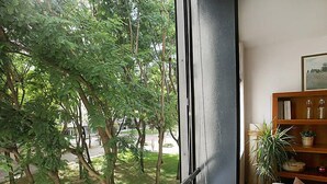 Une pièce dotée d’une grande fenêtre donnant sur un espace verdoyant avec des arbres. On y trouve une bibliothèque en bois remplie de livres et d’objets décoratifs, ainsi qu’une plante en pot posée sur le rebord de la fenêtre.