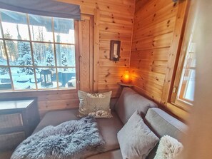 A cozy cabin interior with a sofa, pillows, a fur rug, and a window with a view of a snowy landscape.
