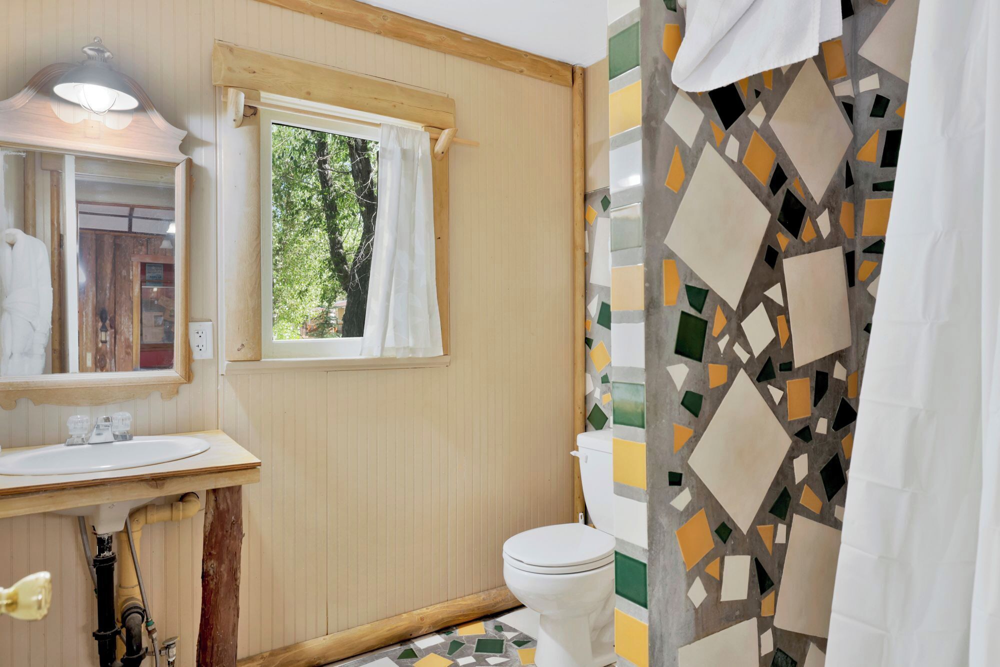 Een badkamer met een houten wastafelmeubel, een spiegel, een raam met uitzicht op bomen, een toilet en een douchegordijn met een geometrisch patroon.