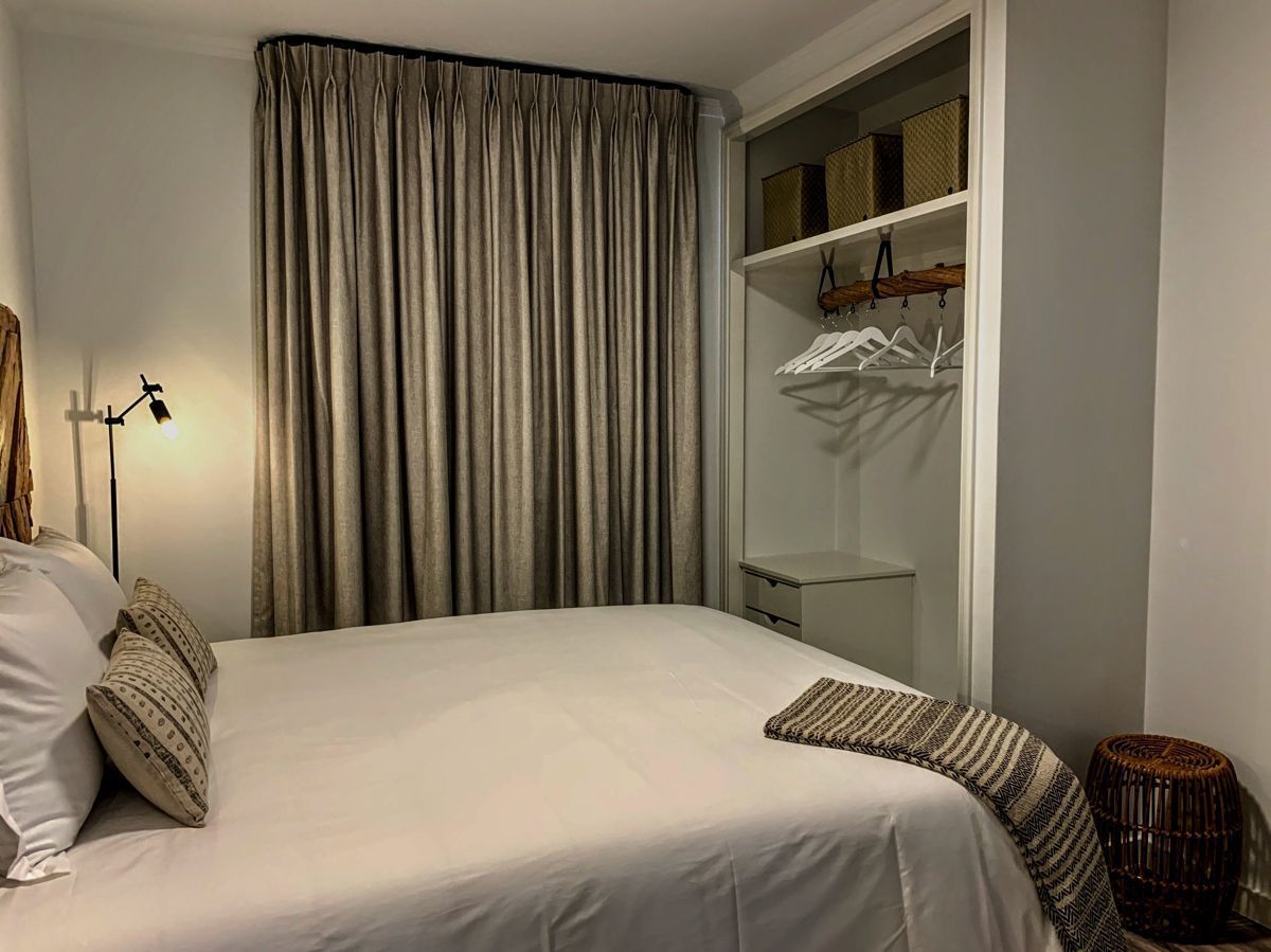 A neatly made bed with pillows, a bedside lamp, a wardrobe with hanging clothes, and a small wicker stool.