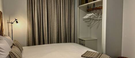 A neatly made bed with pillows, a bedside lamp, a wardrobe with hanging clothes, and a small wicker stool.
