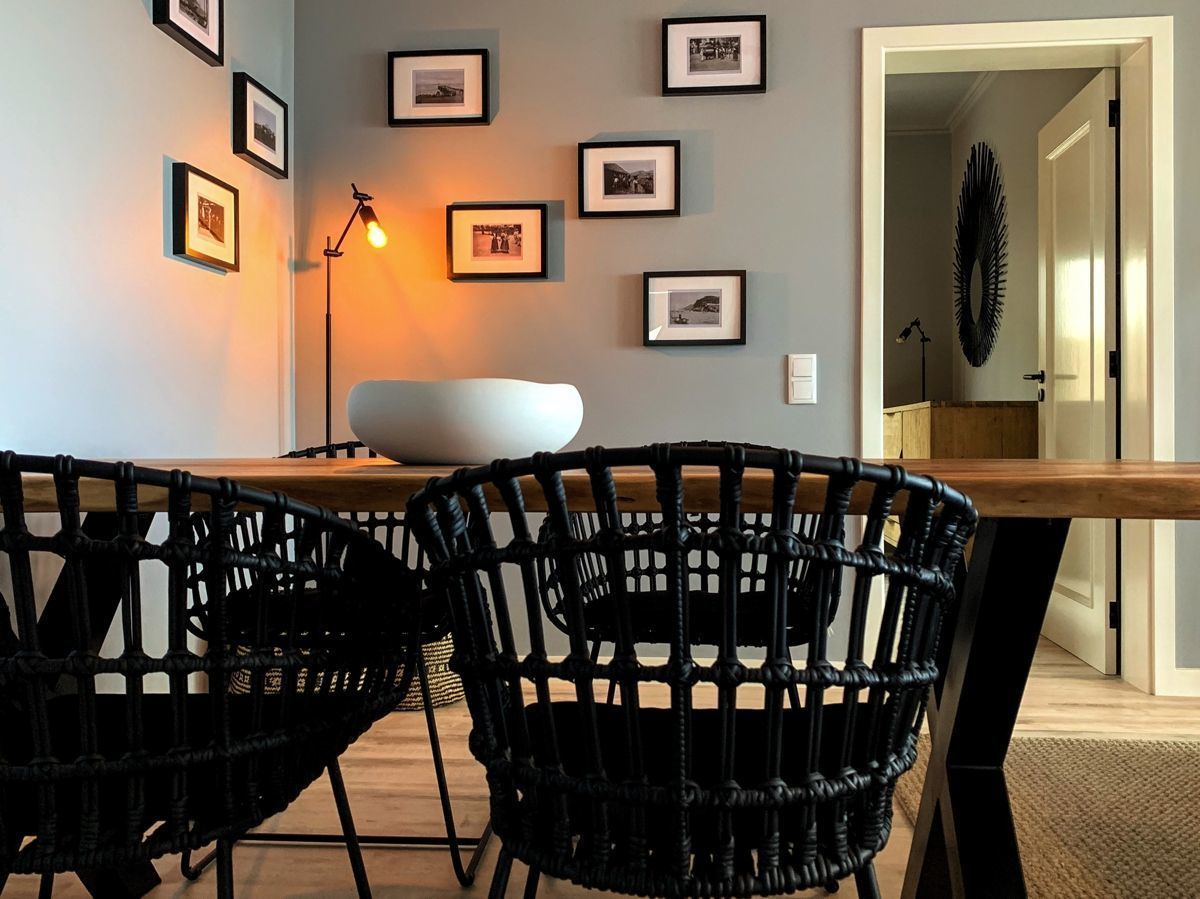 Un coin repas avec une table en bois et des chaises noires en osier, des tableaux encadrés au mur et un lampadaire.