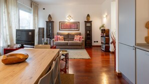 A modern living room with a wooden dining table, a sofa with cushions, a TV, and decorative items.