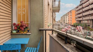 A balcony with a blue table and chairs, a window with flowers, and a view of a city street with buildings.