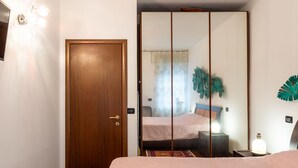 A bedroom with a wooden door, a bed with a pink bedspread, a nightstand with a lamp, and a wall-mounted mirror.
