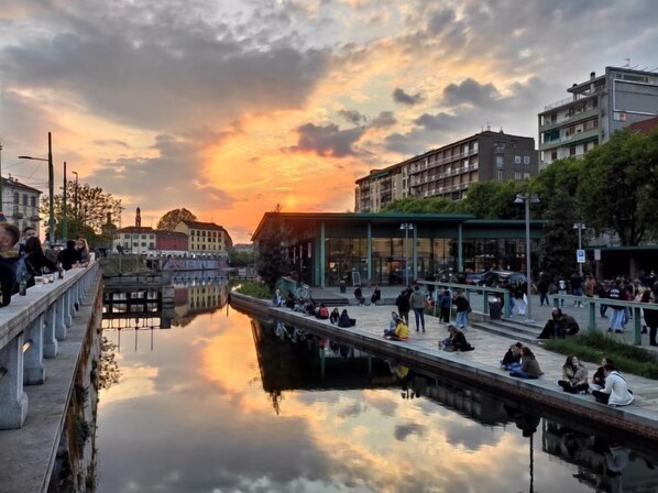 Un paseo fluvial con gente sentada y caminando, rodeado de edificios y un cielo al atardecer.