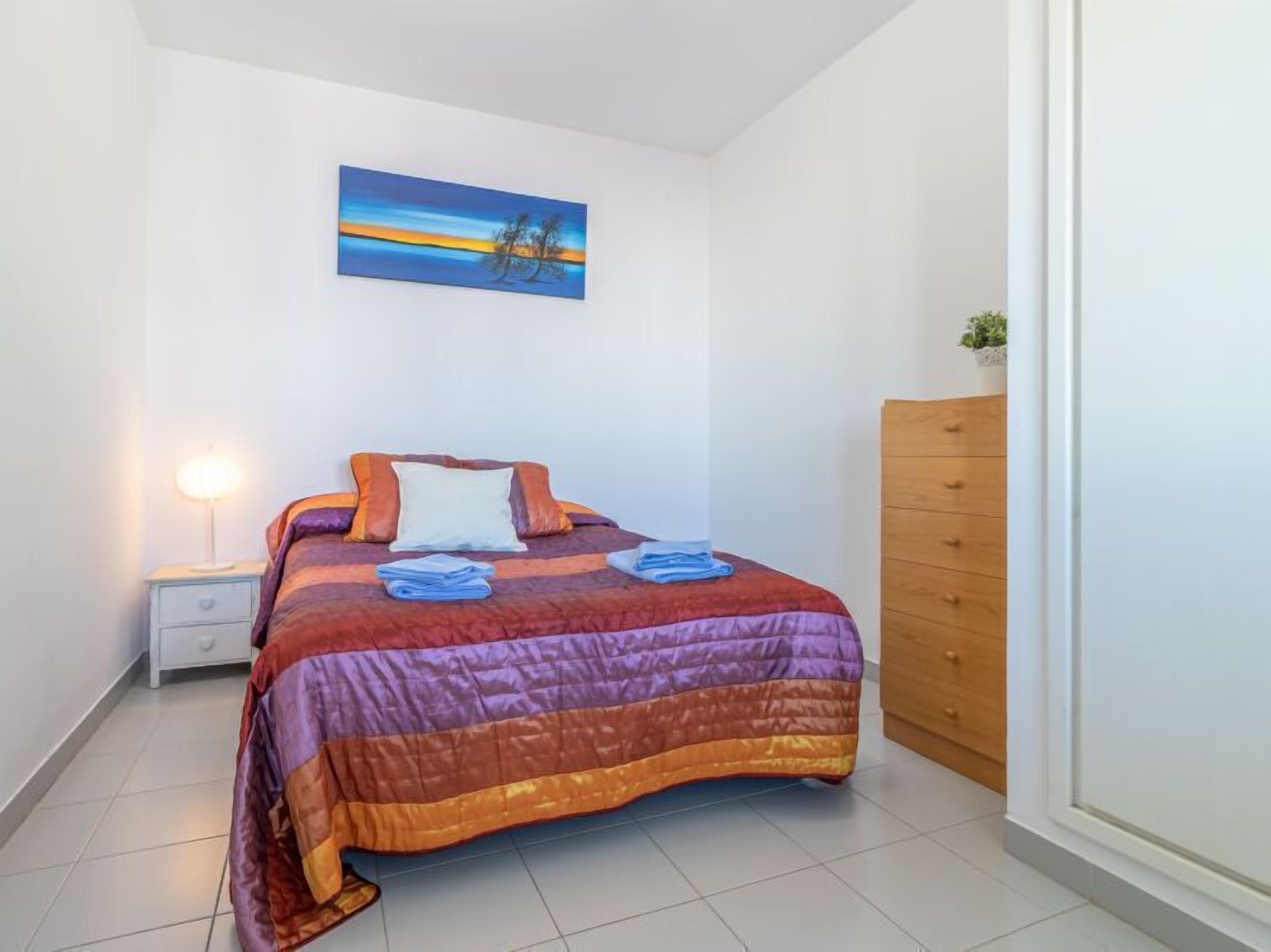A neatly made bed with a colorful quilt, a bedside lamp, and a painting of a sunset on the wall.