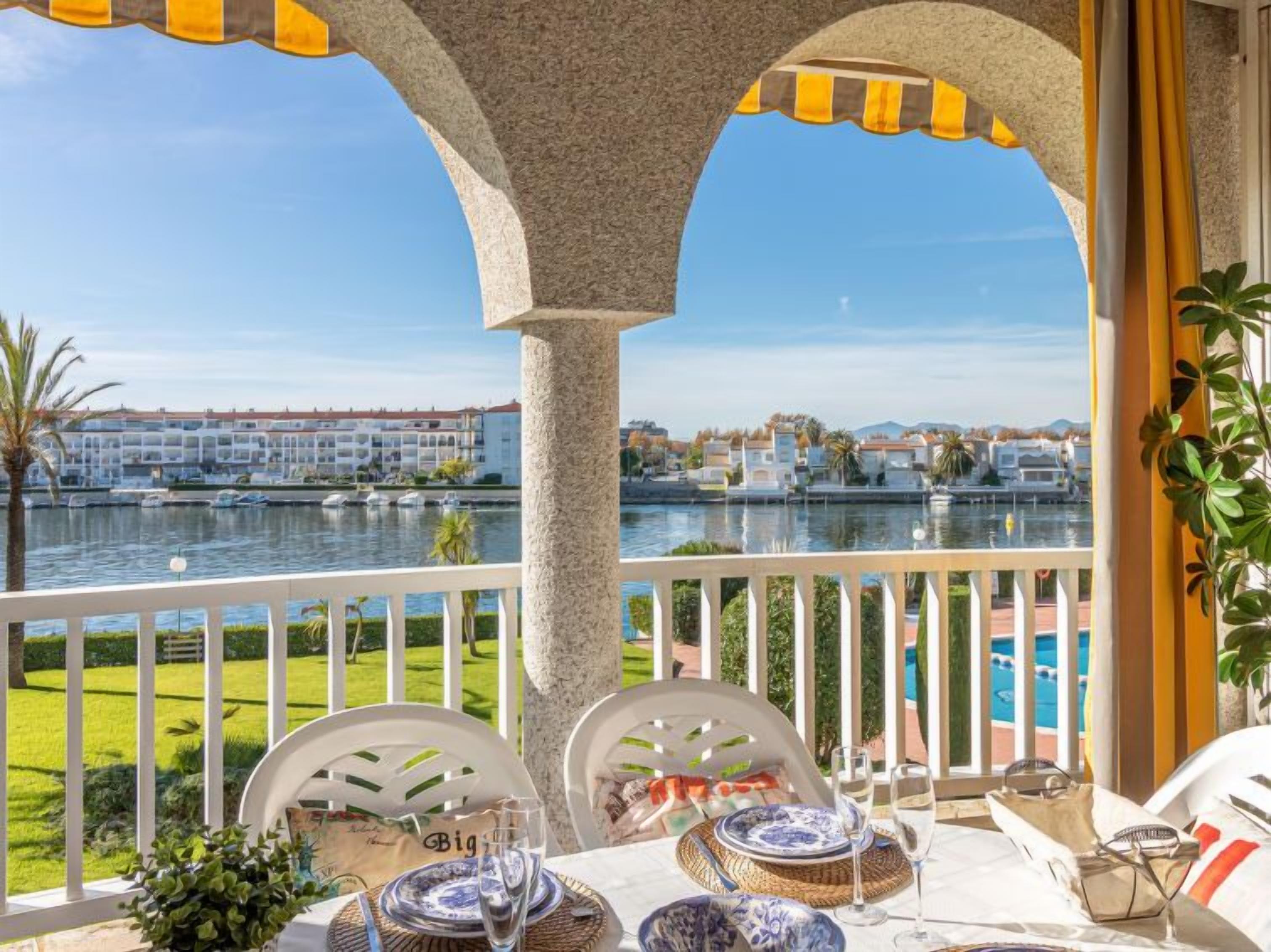 A balcony with a view of a marina and buildings, furnished with white chairs and a table set for two.