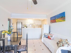 Une cuisine moderne avec des meubles blancs et un îlot central, un coin repas avec une table et des chaises, et un salon avec un canapé et des coussins.