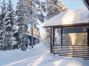 Une cabane en bois recouverte de neige, dotée d’un porche, entourée d’arbres chargés de neige.