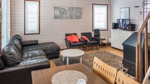 Un vaste espace de vie comprenant un canapé en cuir noir, une table basse ronde et une table à manger en bois. On y trouve deux chaises, un petit tapis et un téléviseur posé sur un meuble. Les murs de la pièce sont en bois et elle donne sur un balcon agrémenté d’une rambarde.