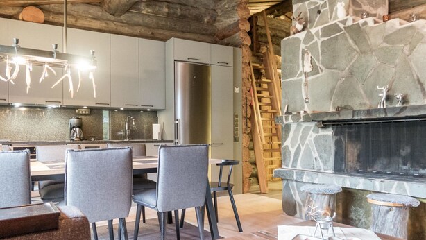 A modern kitchen with a stone fireplace, wooden beams, and a dining area with grey chairs.