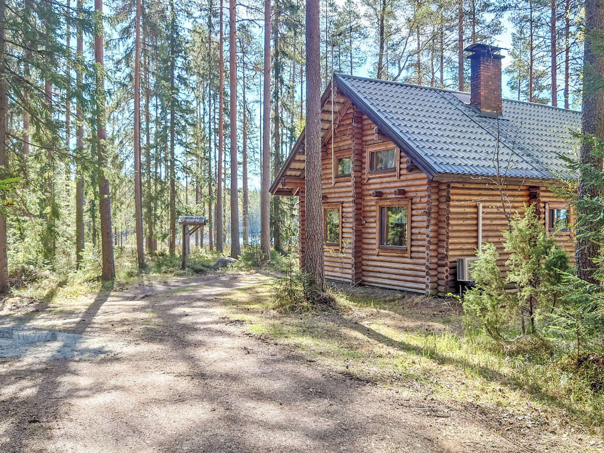 Hirsimökki, jonka päällä on metallikatto, ja sen ympärillä kohoaa korkeita puita. Mökin luo johtaa sorainen polku.