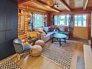 A cozy log cabin interior with a sofa, chairs, a round table, and a large window offering a view of the outdoors. - Kuusela by Interhome (Soini)