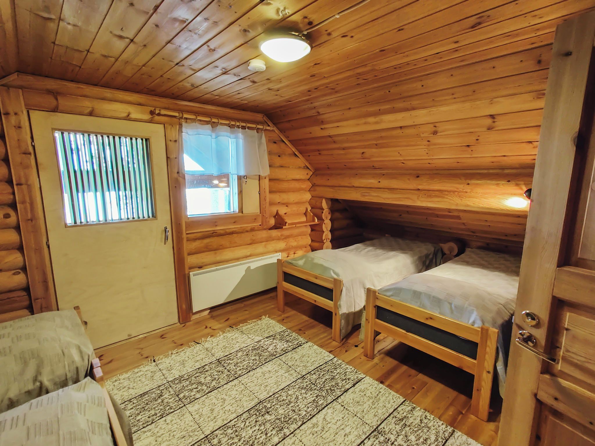 A log cabin bedroom with two beds, a window with blinds, a door, and a ceiling light.