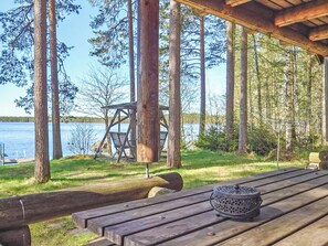 A wooden deck with a bench and a metal lantern, overlooking a lake with trees and a wooden swing set. - Kuusela by Interhome (Soini)