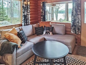 A cozy living room with a sectional sofa, a round coffee table, and a view of a forest through large windows. - Kuusela by Interhome (Soini)