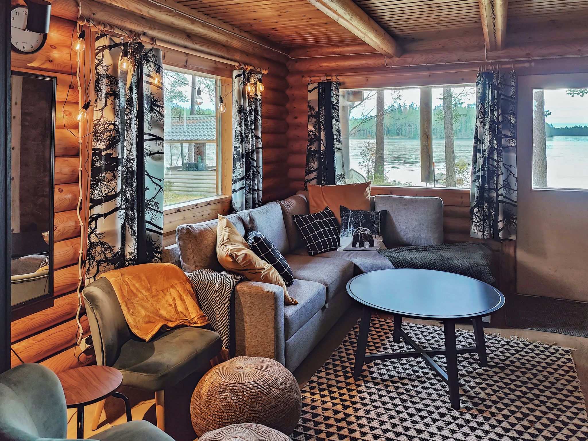 Un interior de cabaña acogedor con un sofá, sillones, una mesa redonda y vistas a un lago a través de grandes ventanales.