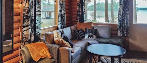 Un interior de cabaña acogedor con un sofá, sillones, una mesa redonda y vistas a un lago a través de grandes ventanales.