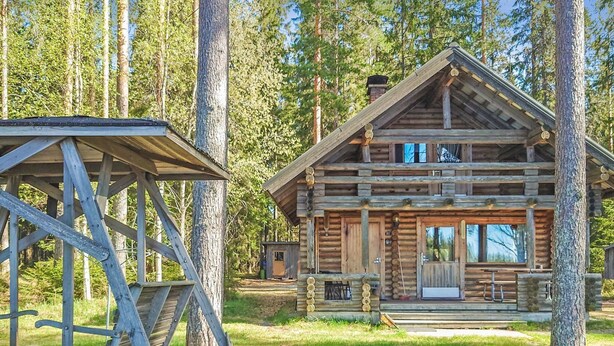 A rustic log cabin with a porch and a wooden deck, surrounded by tall trees.